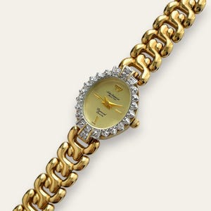 Puede incluir: Reloj de pulsera dorado con esfera ovalada y bisel con diamantes. La esfera es dorada con agujas doradas y un emblema de corona. La correa es de diseño de eslabones. El reloj tiene el texto "Diamond Quartz" y "Since 1993".