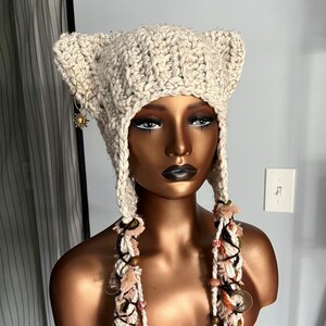 May include: A beige crocheted hat with cat ears and long, beaded tassels. The hat is on a mannequin with a brown head and neck.