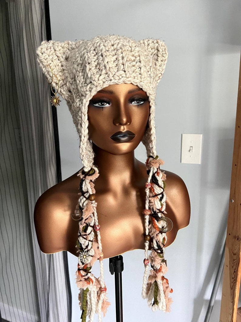 May include: A beige crocheted hat with cat ears and long, beaded tassels. The hat is on a mannequin with a brown head and neck.