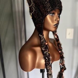 May include: A brown and black crocheted hat with ear flaps and long tassels. The hat has gold hoop earrings attached to the side.