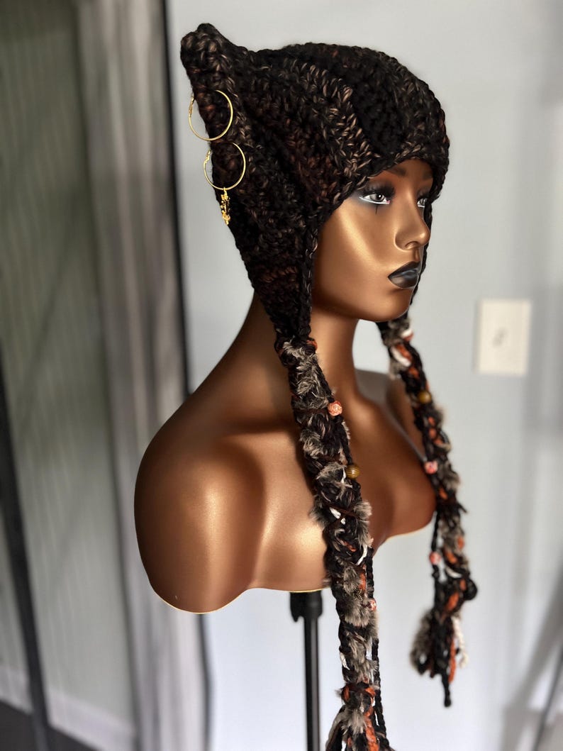May include: A brown and black crocheted hat with ear flaps and long tassels. The hat has gold hoop earrings attached to the side.