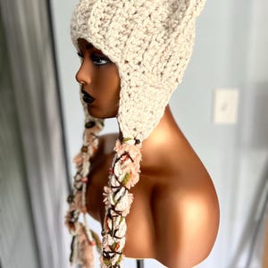 May include: A cream-colored crocheted beanie with ear flaps and long, colorful tassels. The beanie is made with a chunky yarn and has a textured stitch pattern.