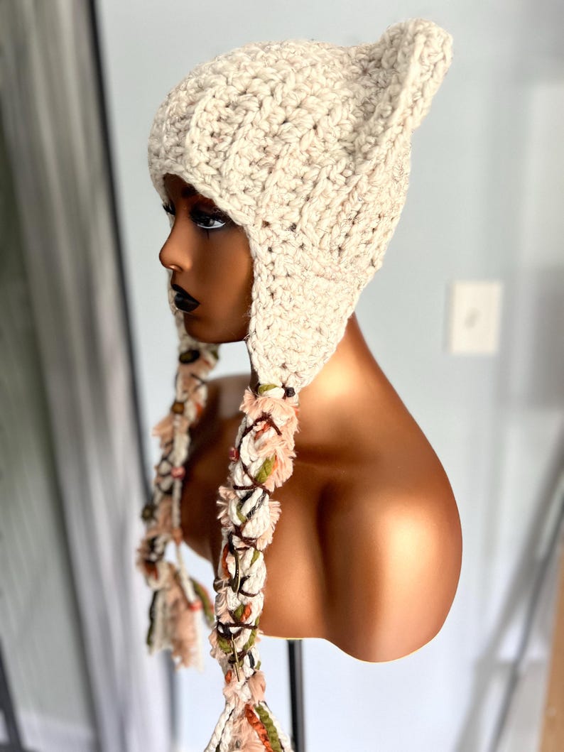 May include: A cream-colored crocheted beanie with ear flaps and long, colorful tassels. The beanie is made with a chunky yarn and has a textured stitch pattern.