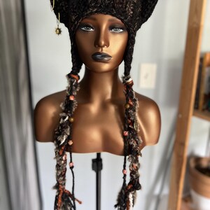 May include: A brown and black crocheted cat ear hat with long, braided tassels featuring beads and charms. The hat is on a mannequin head.