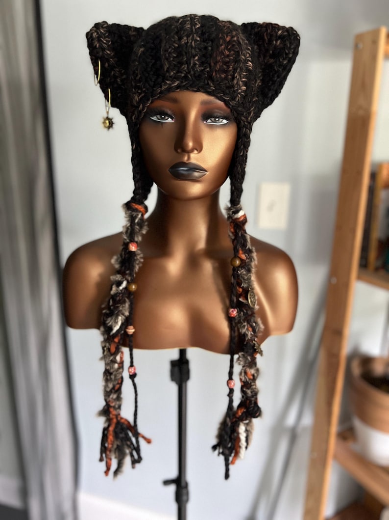 May include: A brown and black crocheted cat ear hat with long, braided tassels featuring beads and charms. The hat is on a mannequin head.