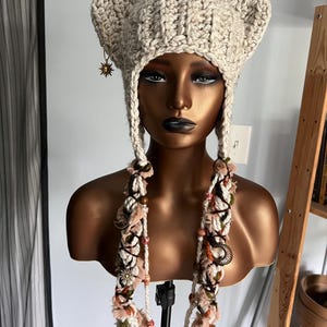May include: A beige crocheted hat with cat ears and long, colorful tassels. The hat is decorated with small, gold-colored charms.