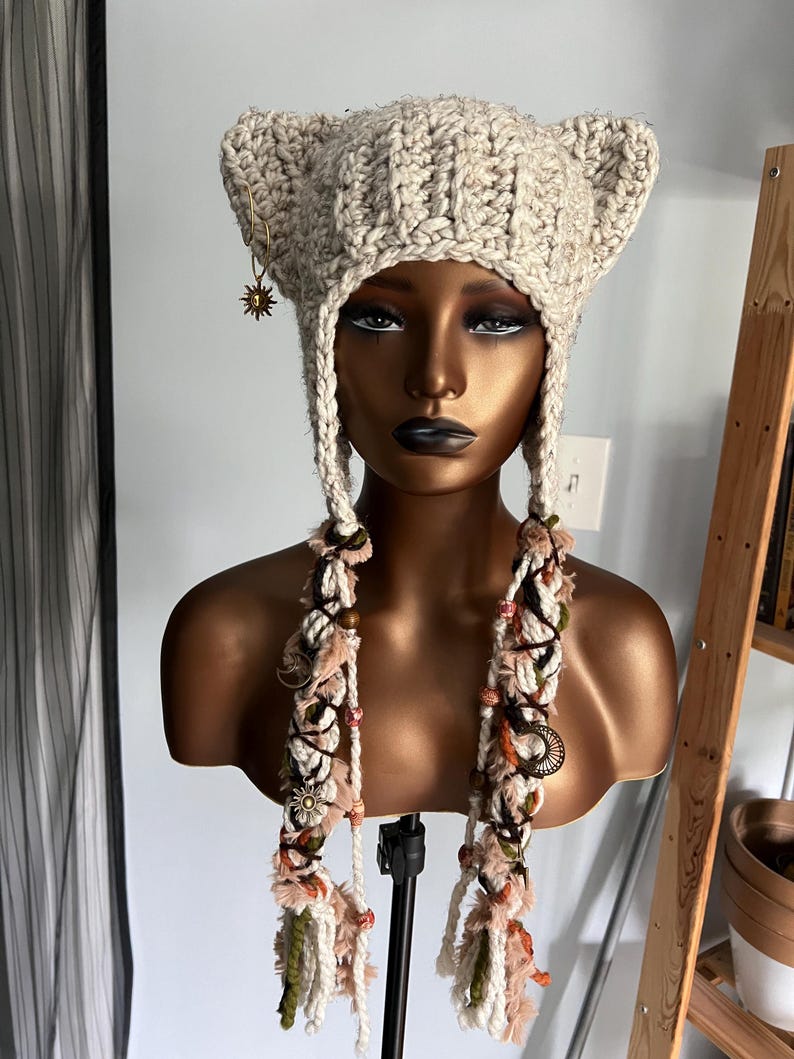 May include: A beige crocheted hat with cat ears and long, colorful tassels. The hat is decorated with small, gold-colored charms.