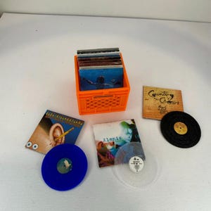 May include: A bright orange plastic crate filled with a stack of vinyl records. Outside the crate are two album covers, one titled Electriclarryland and one titled Alanis, along with a blue vinyl record, a clear vinyl record, and a black record.