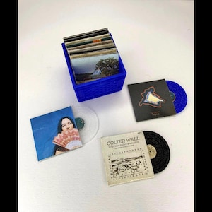 May include: A collection of miniature vinyl records and album covers. A blue plastic crate holds several albums. Individual records include a blue glitter vinyl, a black vinyl, and two with album art featuring a woman and a landscape.
