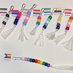 Può includere: Una collezione di portachiavi con perline, chiusure argentate e nappe bianche. Ogni portachiavi presenta un diverso schema di colori che rappresenta varie identità LGBTQ+, tra cui asessuale, bisessuale, transgender, pansessuale, gay, non binario, lesbica e gay/queer.