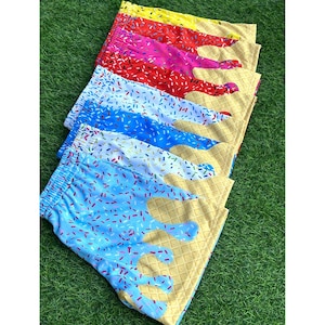 May include: A stack of colorful shorts with an ice cream cone design. The shorts feature a waffle cone base and a variety of ice cream flavors, including yellow, pink, red, blue, and white, with colorful sprinkles. The shorts are displayed on a green surface.