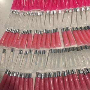 May include: An array of lip gloss tubes with silver lids. The tubes are filled with clear and pink gloss. The arrangement suggests a cosmetic product, with a glossy, smooth finish.