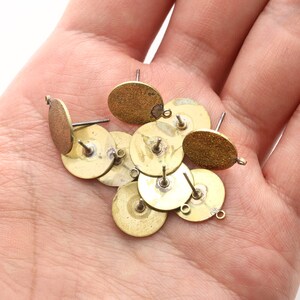 May include: A collection of brass-toned earring posts and oval earring blanks. The earring components have a vintage appearance, with a mix of round and oval shapes. Some have small loops for attaching additional elements.
