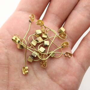 May include: A pile of gold-toned earring findings with a square shape. The findings are made of metal and have a loop at the top for attaching earrings.