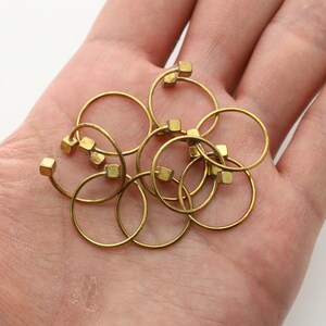 May include: A set of eight gold-toned metal wine glass charms, each featuring a small square charm. The charms are arranged in a pile on a hand.