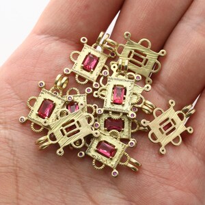 Puede incluir: Un grupo de dijes de metal dorado con piedras preciosas cuadradas de color rosa. Cada dije tiene un diseño detallado con un marco cuadrado y pequeños círculos alrededor de los bordes.