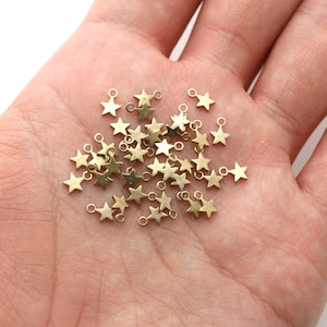 May include: A pile of gold-toned metal star charms with jump rings. The charms are scattered on a person's hand.