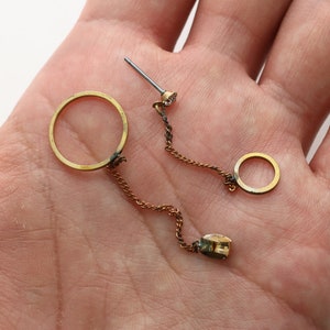 Op de afbeelding: Een paar gouden oorbellen met een ketting die twee ronde hoepels verbindt. Een hoepel is groter dan de andere. De oorbellen hebben een klein, helder steentje als accent.