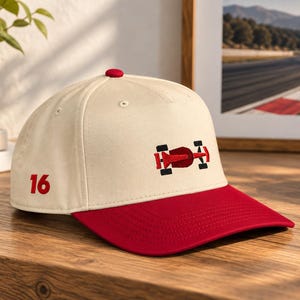Könnte beinhalten: Eine cremefarbene Baseballkappe mit rotem Schirm, die auf der Vorderseite eine gestickte Grafik eines roten und schwarzen Rennwagens sowie auf der Seite die in Rot gestickte Zahl 16 aufweist. Die Kappe hat eine strukturierte Krone und einen roten Knopf.