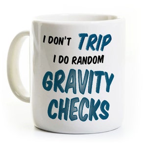 May include: A white ceramic mug with a handle on the left side. The mug features the blue and black text: I DON'T TRIP I DO RANDOM GRAVITY CHECKS. The words TRIP, GRAVITY, and CHECKS are in a bold blue font, while I DON'T and I DO RANDOM are black.