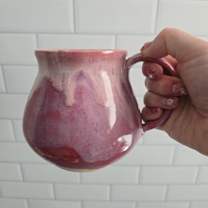 Peut inclure: Une tasse en céramique arrondie avec un vernis brillant présentant un dégradé de magenta profond et de violet à la base, passant à un motif de coulures rose crème plus clair près du bord. La tasse possède une anse incurvée et une surface lisse.