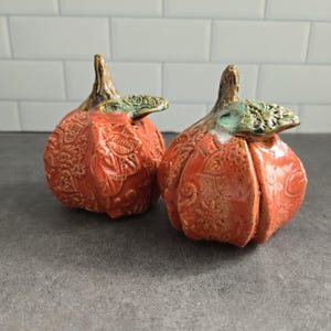 Peut inclure: Deux figurines de citrouilles en céramique avec un vernis orange brillant et des motifs floraux en relief complexes. Chaque citrouille présente une tige brune texturée et une feuille verte attachée au sommet, avec un aspect artisanal.
