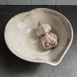 May include: A ceramic grater bowl with an irregular, organic rim and a glossy, cream-coloured glaze featuring soft purple and pink highlights. The centre of the bowl has a textured, raised surface for grating, and two whole garlic bulbs rest inside.