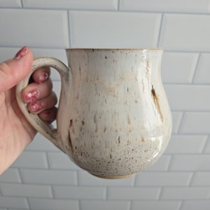 Peut inclure: Une tasse en céramique avec un corps arrondi et une anse incurvée, présentant un émail blanc cassé avec des taches brunes et des stries verticales. La surface a un aspect texturé avec des points brun foncé concentrés près de la base.