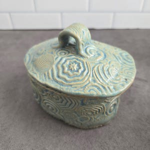 May include: An oval ceramic lidded box in a light blue and cream glaze. The surface features raised, textured patterns of concentric circles, spirals, and geometric shapes. A curved handle sits atop the lid, matching the overall textured design.
