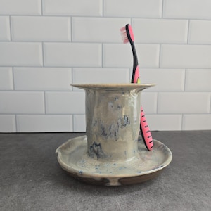May include: A ceramic toothbrush holder with a wide, flat base and a cylindrical centre, featuring a mottled beige and blue glaze. A single toothbrush with a black handle and pink bristles is inserted into the holder, resting against the side.