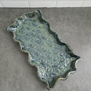 Peut inclure: Un plateau rectangulaire en céramique avec un bord ondulé et festonné, recouvert d'une glaçure brillante bleu-vert marbrée. La surface présente un motif en relief de tourbillons répétés, créant un aspect texturé avec des nuances variées.