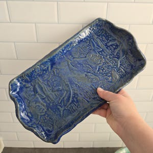 May include: A rectangular ceramic tray with a deep blue glaze and an embossed underwater scene featuring whales, fish, and coral textures. The tray has an irregular, wavy edge and a glossy finish, showcasing intricate relief details across the surface.