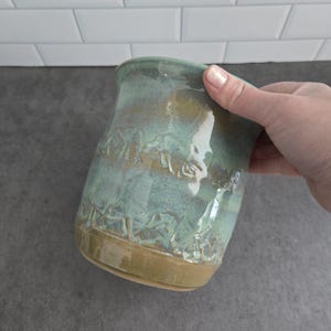 May include: A ceramic mug featuring a glossy glaze in shades of sage green and teal. The exterior surface is decorated with a repeating embossed pattern of mushrooms encircling the body. The base of the mug is a solid, unglazed tan clay colour.