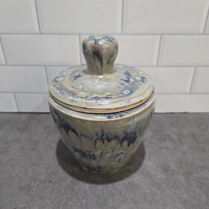 May include: A ceramic lidded jar featuring a mottled glaze in shades of blue, beige, and grey. The jar has a rounded body and a matching lid with a central knob handle. The surface displays a fluid, dripped pattern of colours throughout the piece.