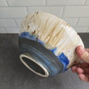 Peut inclure: Un bol en céramique présentant une base gris anthracite qui se transforme en une bande bleue vive, surmontée d'un émail de couleur crème qui coule vers le bas. Le bol a une forme arrondie lisse avec un pied distinct et une finition brillante.