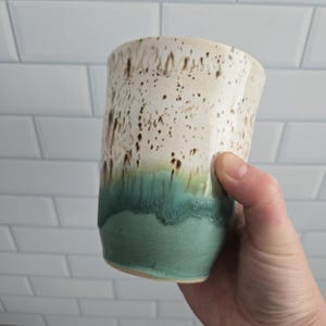Peut inclure: Un gobelet en céramique présentant un design à deux tons. La partie supérieure est de couleur blanc cassé avec des taches brun foncé, tandis que la section inférieure est d'un vert sarcelle mat uni. La transition montre un effet de coulure.