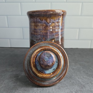 May include: A cylindrical ceramic canister with a matching lid, featuring a glossy glaze in shades of deep brown, amber, and hints of blue. The lid has a raised circular knob in the centre, and the body displays a smooth, reflective surface.