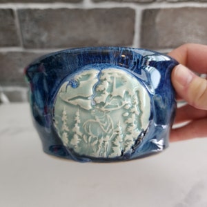 Peut inclure: Un bol en céramique fait à la main avec un glaçage bleu foncé. Le bol présente un motif en relief d'un cerf dans une scène de forêt, avec des accents bleu clair et blancs. Le bol est tenu par une main.