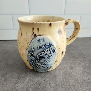 May include: A ceramic mug with a speckled yellow glaze and a blue and white bird design. The mug has a rounded body and a handle.