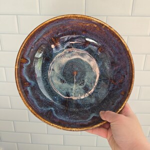 Peut inclure: Un bol en céramique rond présentant un vernis brillant avec des motifs tourbillonnants dans des tons bleu marine profond, violet foncé et brun terreux. Le centre du bol affiche une zone circulaire plus claire, bleu et gris moucheté.