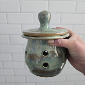 May include: A round ceramic garlic keeper with a lid, featuring a variegated glaze in shades of sage green, blue, and brown. The container has a wide rim, a knob-style handle on the lid, and several circular ventilation holes on the body.