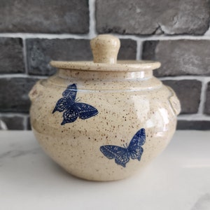 May include: A beige ceramic jar with a lid, decorated with two blue butterfly motifs. The jar has a speckled texture and small handles. The lid features a small knob. A decorative storage container, ideal for the kitchen.