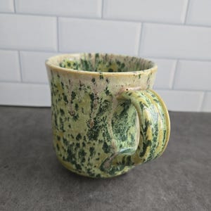 Peut inclure: Une tasse en céramique avec un émail tacheté dans des tons de vert clair, vert foncé et crème. La tasse présente un corps arrondi et une anse épaisse et incurvée avec un motif moucheté assorti, offrant une finition texturée sur toute la surface.