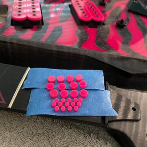 May include: Close-up of a pink and black electric guitar with a zebra print design. The guitar has two black knobs and two black pickup covers. The guitar is on a blue surface with a collection of pink dots.