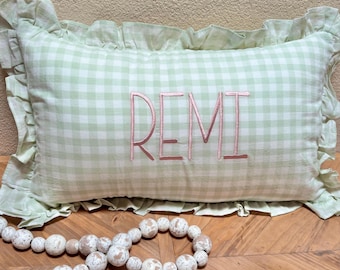 May include: A rectangular pillow with a light green and white gingham pattern. The word "REMI" is embroidered in pink thread across the center. The pillow has a ruffled edge and is resting on a wooden surface.