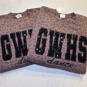 May include: Two mauve-colored sweatshirts with a leopard print pattern. Each sweatshirt has the letters "GWHS" in black sequins, with the word "dance" embroidered below. The sweatshirts are likely for a school dance team.