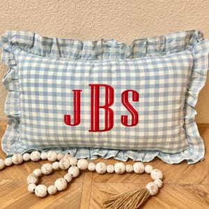 May include: A rectangular pillow with a light blue and white gingham pattern and a ruffled edge. The pillow features the embroidered initials "JBS" in red. A decorative beaded garland and tassel are in the foreground.