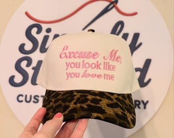 May include: A white baseball cap with a leopard print brim. The cap features pink embroidered text that reads "Excuse Me, you look like you love me". The cap is held in front of a white sign with the words "Simply Stitched".