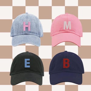 May include: Four baseball caps in different colors: light blue with a pink "H", pink with a white "M", black with a blue "E", and navy blue with a red "B". The caps are arranged against a checkered background.