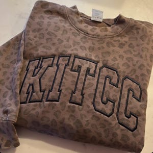 May include: A brown sweatshirt with a leopard print pattern. The word "KITCC" is embroidered across the front in a dark blue, block letter style. The sweatshirt is folded on a white surface.